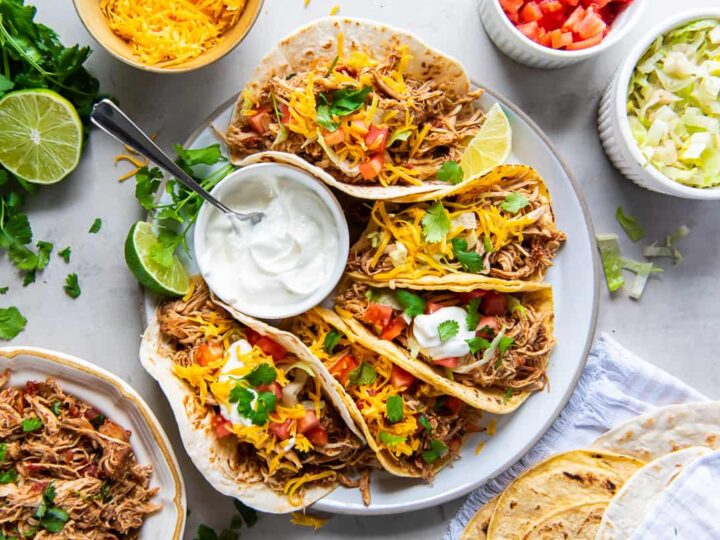 A plate of soft shell tacos filled with shredded chicken, cheese, and diced tomatoes, garnished with cilantro. A small bowl of sour cream with a spoon is placed in the center. Sides of lime, cilantro, shredded cheese, and cabbage surround the plate.