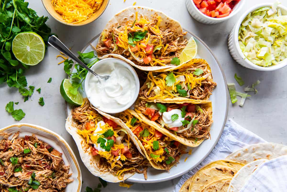 A plate of soft shell tacos filled with shredded chicken, cheese, and diced tomatoes, garnished with cilantro. A small bowl of sour cream with a spoon is placed in the center. Sides of lime, cilantro, shredded cheese, and cabbage surround the plate.