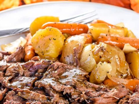A plate of tender pot roast with gravy, accompanied by carrots, potatoes, and onions. The background features vibrant autumn leaves, adding a seasonal touch. A fork is placed on the plate, ready for use.