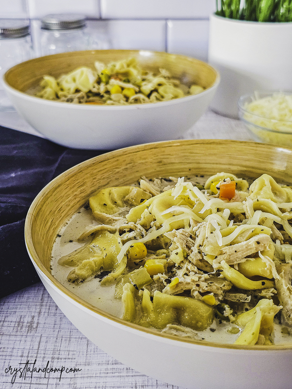 Two bowls of creamy chicken tortellini soup garnished with shredded cheese, with visible carrots and herbs. A black napkin and a small bowl of extra cheese are on the side. A potted plant decorates the background.
