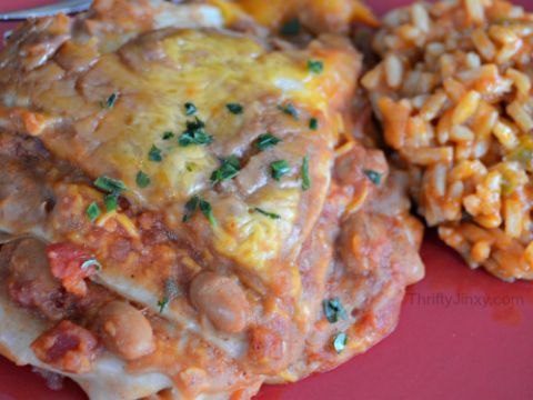 A close-up of enchiladas smothered in sauce and cheese, garnished with herbs. They are served on a red plate with a side of Mexican-style rice. A fork is partially visible in the background.