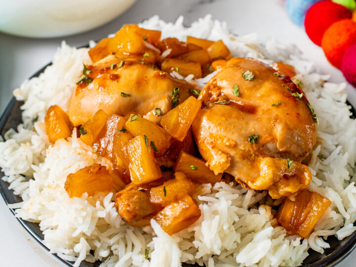 A plate of white rice topped with glazed chicken and chunks of pineapple. The dish is garnished with herbs. In the background, there are bowls of rice and colorful decoration balls. A fork is placed next to the plate.