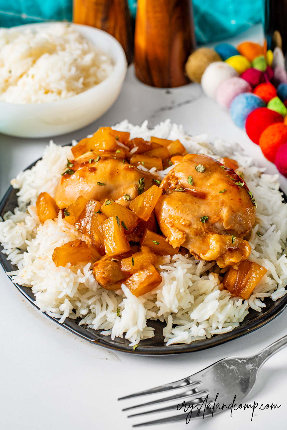 A plate of white rice topped with glazed chicken and chunks of pineapple. The dish is garnished with herbs. In the background, there are bowls of rice and colorful decoration balls. A fork is placed next to the plate.