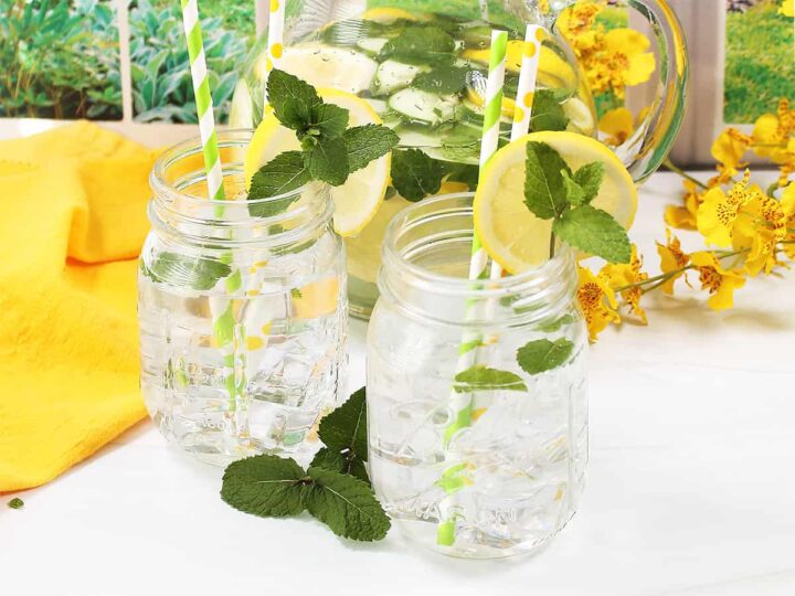 Two mason jars filled with ice water, garnished with lemon slices, mint leaves, and striped straws. A jug of lemon-mint water is in the background, along with yellow flowers and a yellow cloth. The setting is bright and summery.