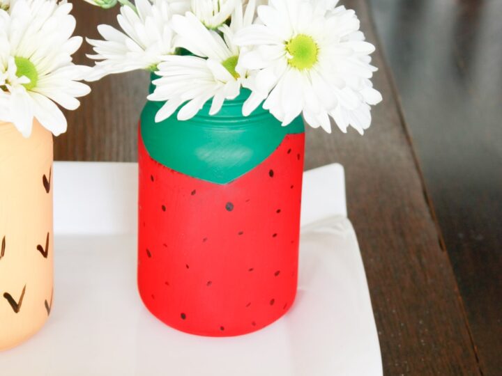 A decorative jar painted to resemble a strawberry, filled with white daisies, sits on a white rectangular tray. The jar features a green top with black seeds. A wooden table and white chair are visible in the background.