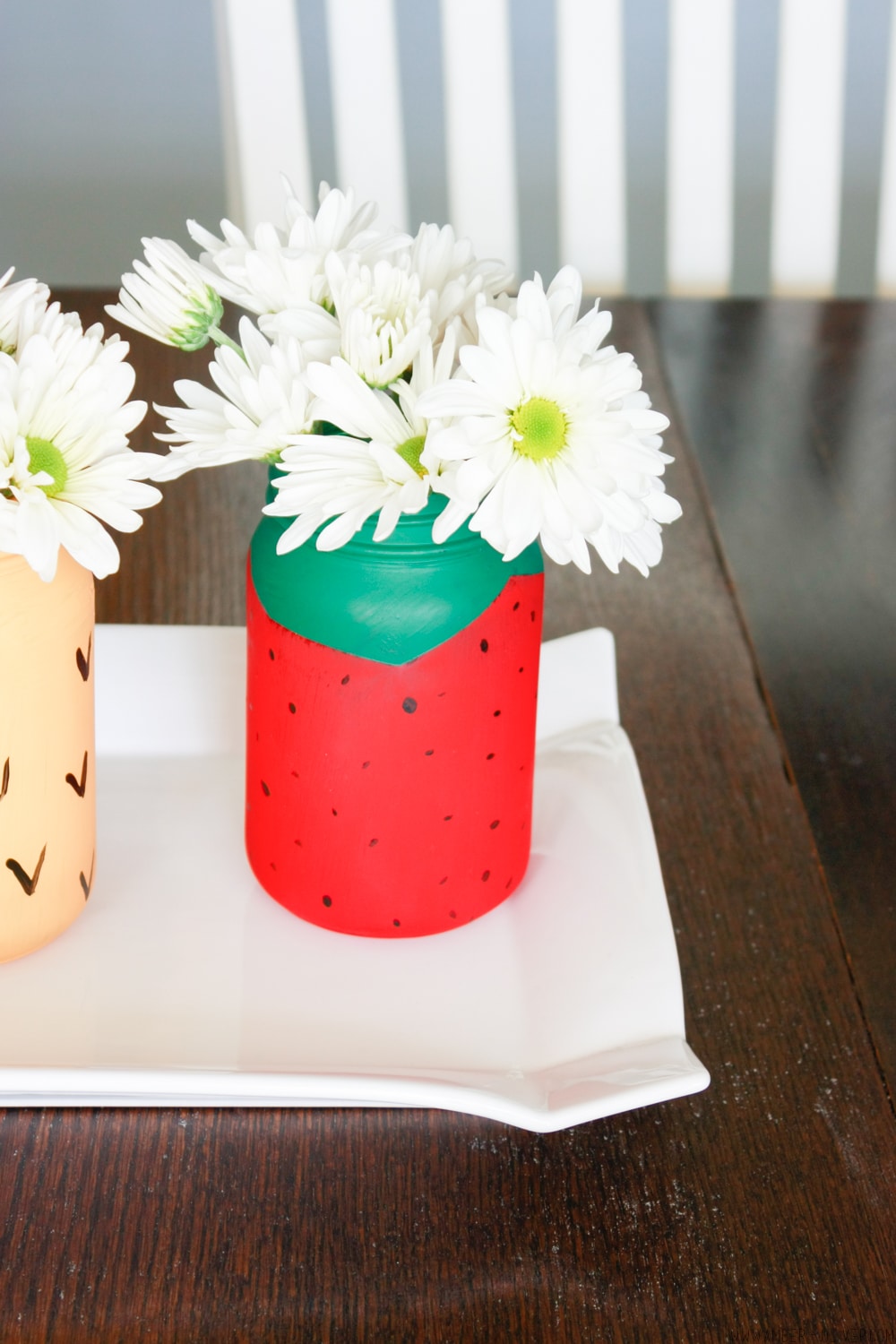 A decorative jar painted to resemble a strawberry, filled with white daisies, sits on a white rectangular tray. The jar features a green top with black seeds. A wooden table and white chair are visible in the background.