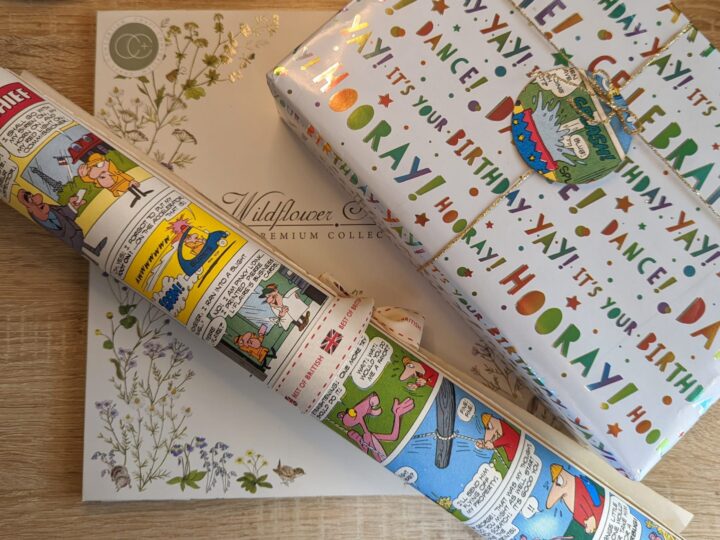 A rolled-up comic strip-themed gift wrap lies atop a floral Wildflower Collection box and a colorful wrapped gift with Hooray! Its your birthday! text. A small gift tag featuring a polar bear is attached to the package.