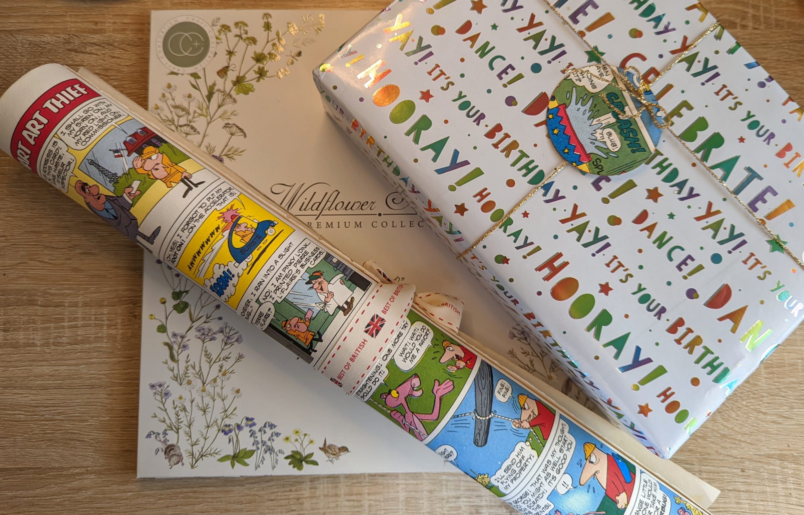 A rolled-up comic strip-themed gift wrap lies atop a floral Wildflower Collection box and a colorful wrapped gift with Hooray! Its your birthday! text. A small gift tag featuring a polar bear is attached to the package.