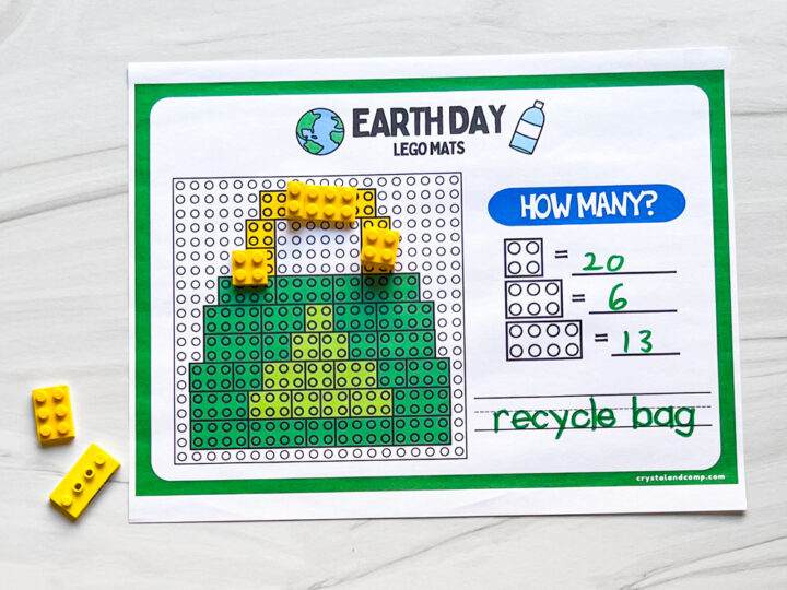 A worksheet titled Earth Day Lego Mats features a pixel art recycle bag design made with green and yellow Lego bricks. Beside the design are math questions: 20 - 6 = 13. Loose yellow bricks are next to the sheet.
