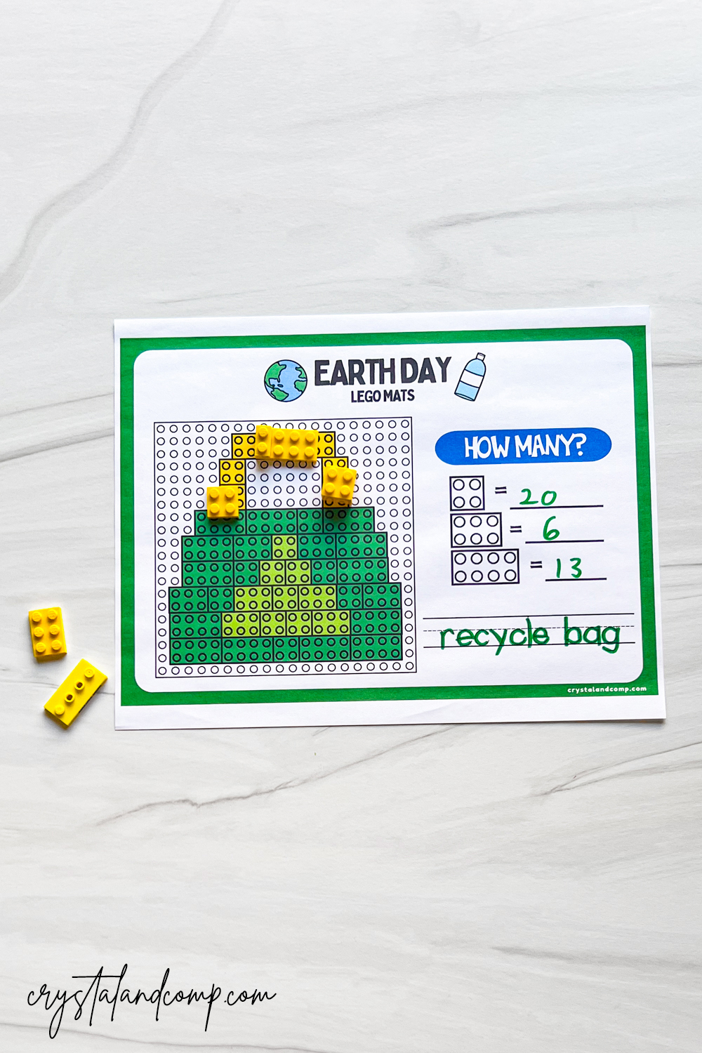 A worksheet titled Earth Day Lego Mats features a pixel art recycle bag design made with green and yellow Lego bricks. Beside the design are math questions: 20 - 6 = 13. Loose yellow bricks are next to the sheet.