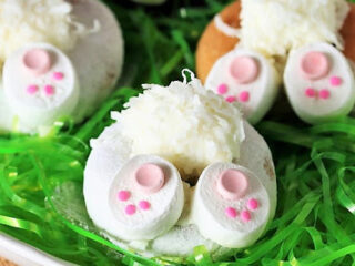 Donuts decorated to resemble bunny bottoms with white frosting tails and pink paw marshmallows rest on green shredded paper in a white plate.