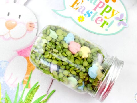 A glass jar filled with green split peas and pastel-colored rabbit-shaped candies lies on its side. Nearby, a decorative Happy Easter! sign and a smiling bunny graphic add a festive touch.