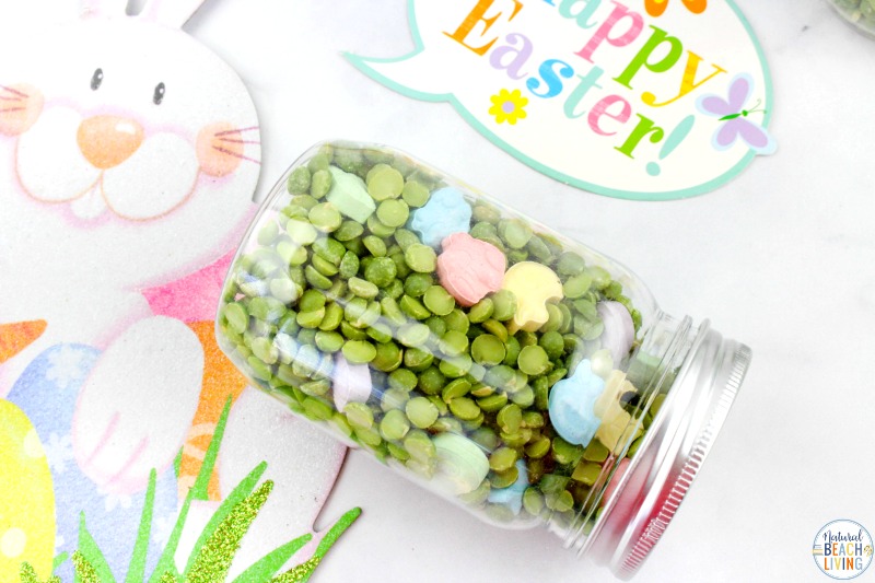 A glass jar filled with green split peas and pastel-colored rabbit-shaped candies lies on its side. Nearby, a decorative Happy Easter! sign and a smiling bunny graphic add a festive touch.