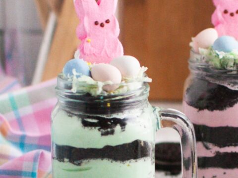 A jar filled with layered mint-green and chocolate dessert, topped with a pink marshmallow bunny and pastel candy eggs. Another similar jar is partially visible next to it. A colorful striped cloth is in the background.