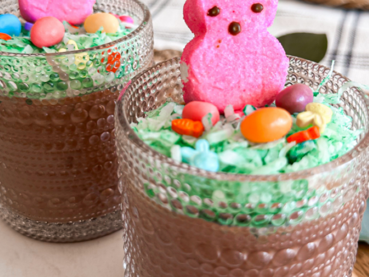Two glass cups filled with chocolate mousse, topped with green coconut shreds, colorful candy eggs, and pink marshmallow bunny peeps. The background shows a textured fabric and a wooden surface.