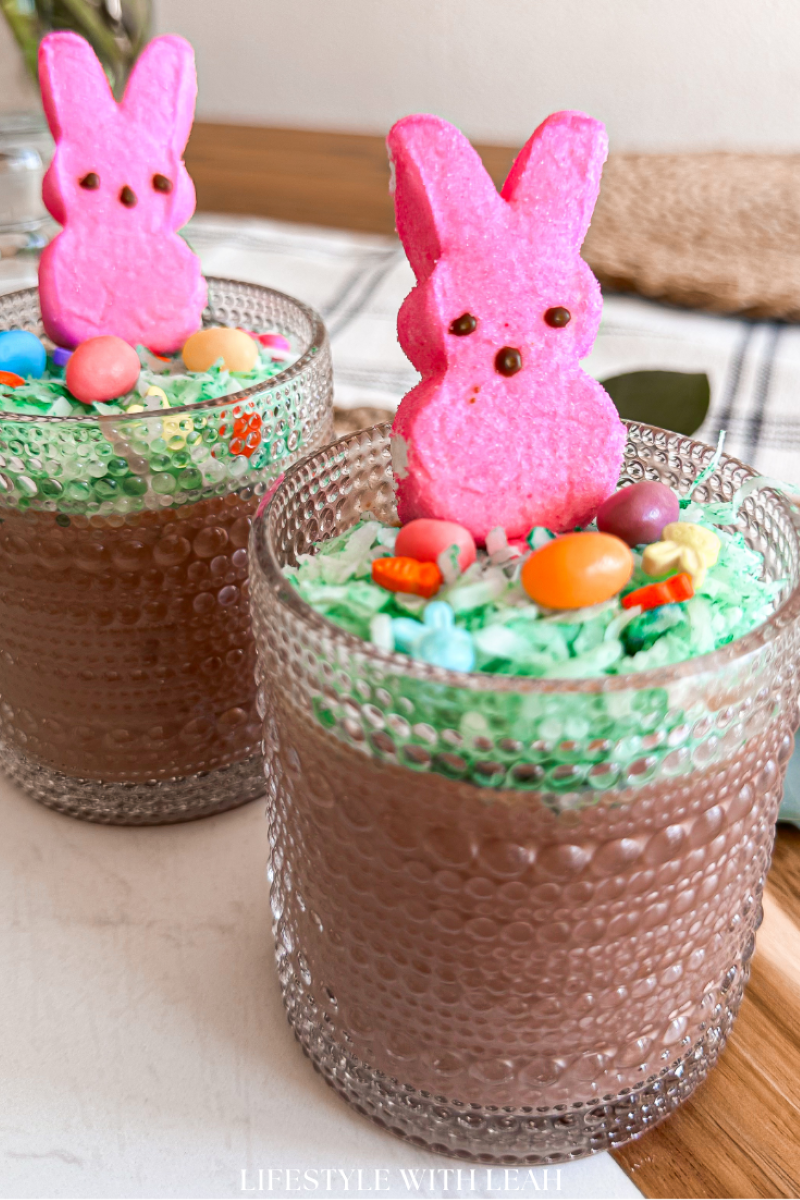 Two glass cups filled with chocolate mousse, topped with green coconut shreds, colorful candy eggs, and pink marshmallow bunny peeps. The background shows a textured fabric and a wooden surface.
