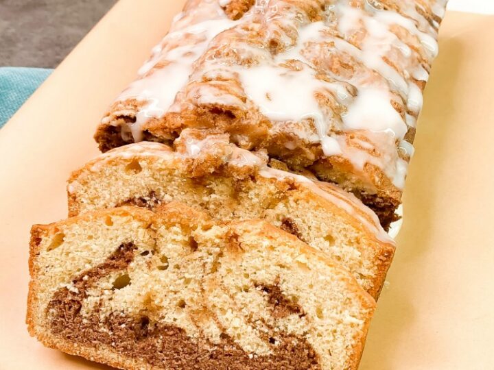 A loaf of glazed cinnamon swirl bread with two slices cut and visible. The bread shows a marbled pattern of cinnamon and plain dough, topped with a light glaze, on a parchment-lined surface.