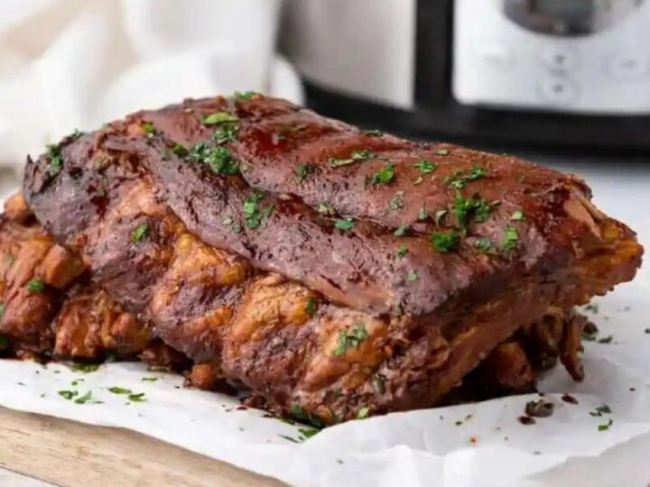Cooked ribs garnished with chopped herbs, placed on a wooden board with parchment paper. A slow cooker is visible in the background.