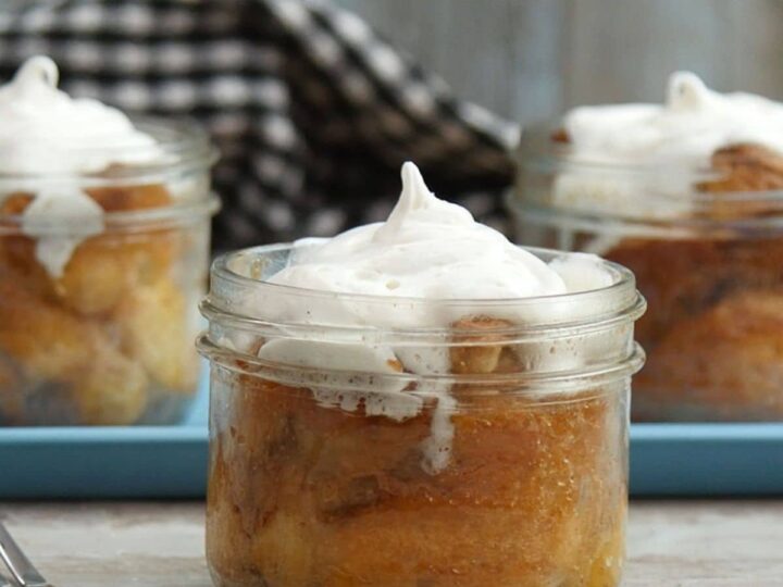 Three glass jars filled with bread pudding topped with whipped cream are placed on a wooden table. A checkered cloth is in the background.