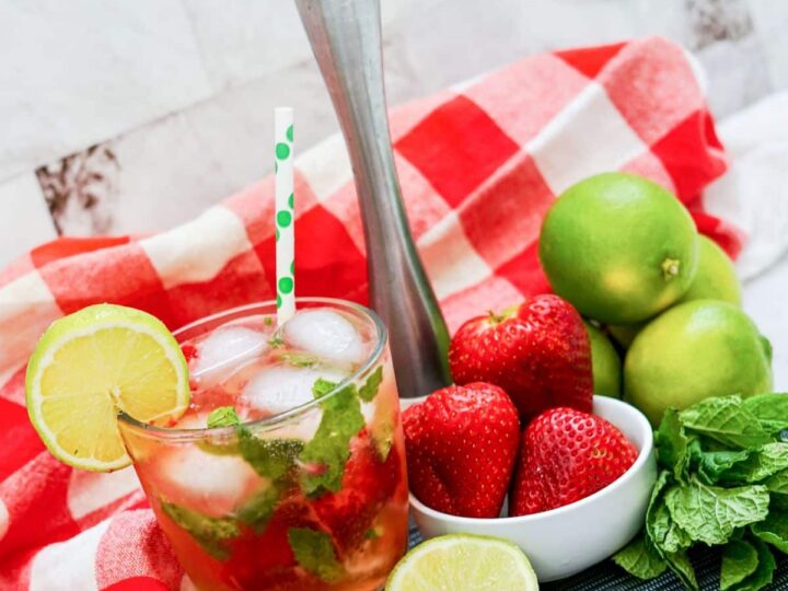 A strawberry mojito garnished with a lime slice and a polka dot straw sits on a red and white checkered cloth. Fresh strawberries, limes, and mint leaves are placed nearby, along with a muddler.
