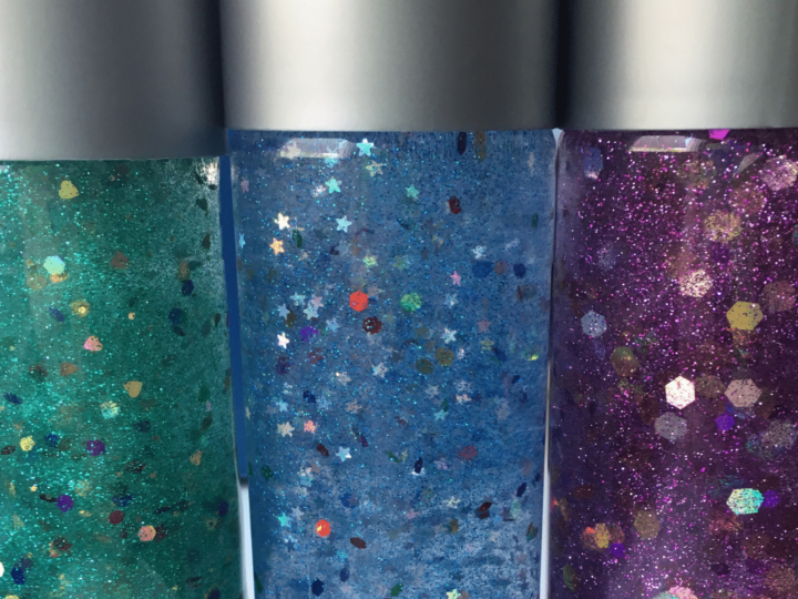 Three glittery sensory bottles. The left bottle is teal with fine glitter, the middle is blue with star-shaped confetti, and the right is purple with hexagonal glitter. All bottles have silver caps and are filled with suspended particles.