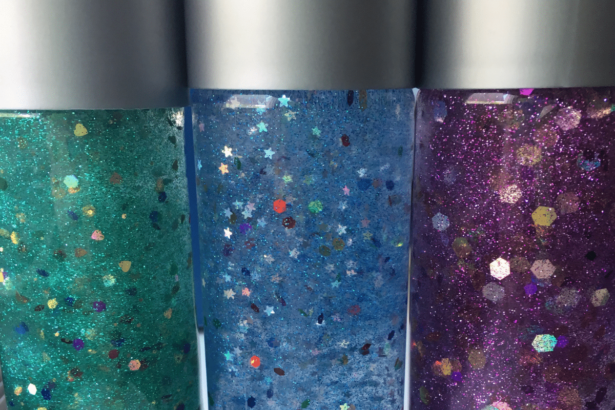 Three glittery sensory bottles. The left bottle is teal with fine glitter, the middle is blue with star-shaped confetti, and the right is purple with hexagonal glitter. All bottles have silver caps and are filled with suspended particles.