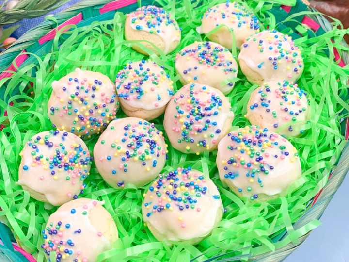 A woven basket with green shredded paper holds a dozen frosted cookies. Each cookie is topped with white icing and colorful sprinkles, creating a festive appearance.