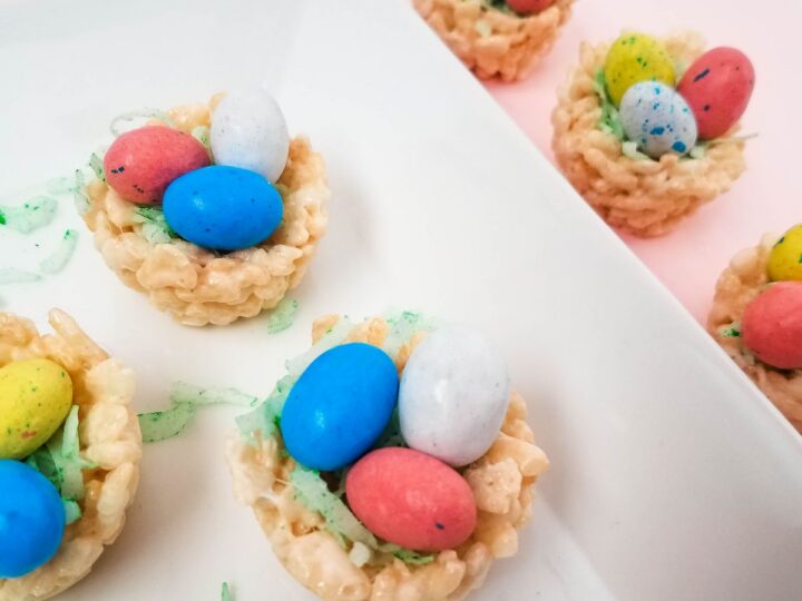 Small Rice Krispie nests holding colorful candy eggs are arranged on a white plate. Green coconut shreds mimic grass, adding a festive touch. The background is a soft pink hue, enhancing the playful, spring-like atmosphere.