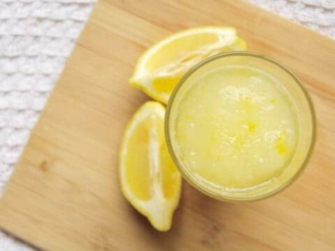 A glass filled with lemon sorbet is placed on a wooden cutting board, next to three lemon wedges. The background features a textured white surface.