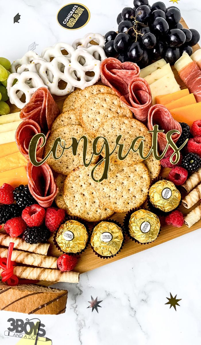 A celebratory charcuterie board perfect for a graduation, featuring assorted cheeses, meats, fruits, pretzels, crackers, and chocolates. Congrats is elegantly scripted across the center. A small 3 Boys and a Dog logo completes this beautifully crafted centerpiece.
