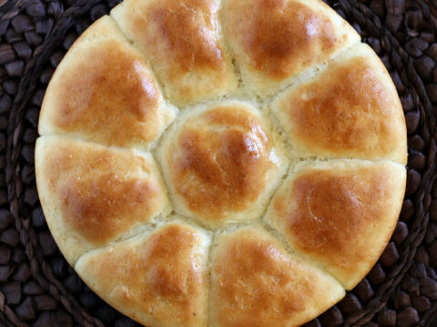 A round loaf of gluten-free bread with a golden-brown crust, divided into eight sections, rests on woven, dark placemats.