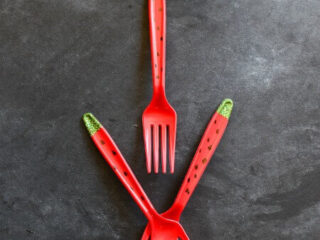Three red plastic forks designed to resemble watermelon slices are arranged on a dark gray surface. Each fork features green tips and black seeds painted on the handles.