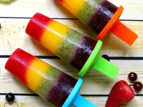 Three colorful rainbow fruit popsicles on wooden boards, each with a layer of red, orange, yellow, green, and purple, in blue, orange, and green plastic molds. Surrounded by assorted fruits like kiwi, strawberries, and berries. Text: Rainbow fruit popsicles.
