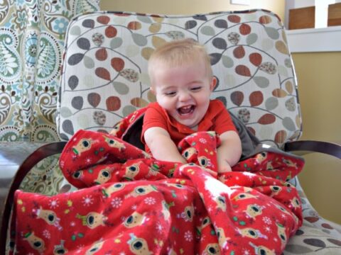 A smiling toddler in a red shirt sits on a patterned armchair, wrapped in a red blanket with floral designs. Sunlight filters in, creating a warm atmosphere. A framed letter W is on the wall behind.