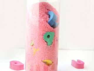 A clear plastic bottle filled with pink sand and colorful foam letters and numbers. Several foam pieces, including letters and numbers, are scattered around the bottle. The text at the top reads I Spy ABC Bottle.