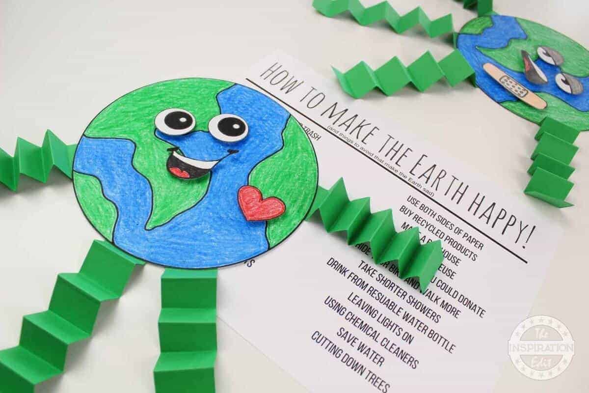 Craft project showing a smiling Earth with googly eyes, a red heart sticker, and accordion-folded green legs. Background shows instructions titled How to Make the Earth Happy with tips for recycling and saving energy.