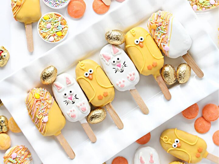 A display of colorful cake pops decorated as animals, including cats and birds, on sticks. They are surrounded by small bowls of sprinkles and golden eggs on a white tray.