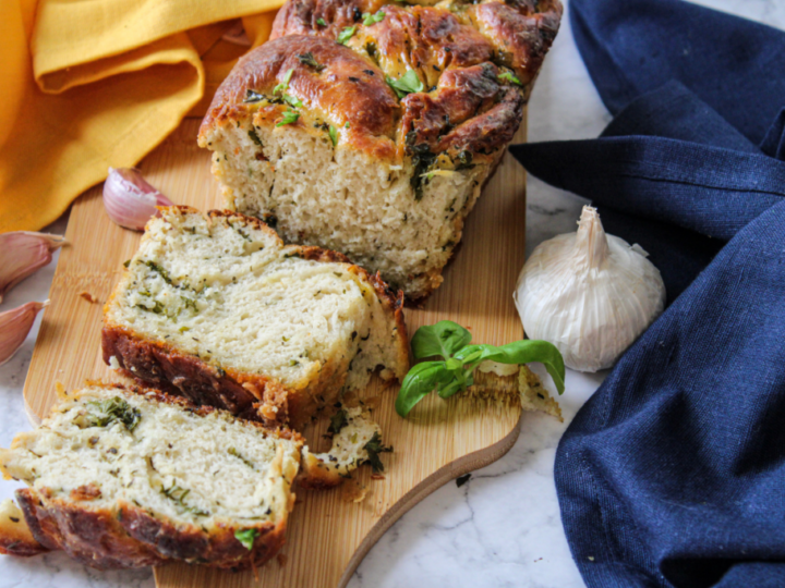 A loaf of savory herb bread on a wooden cutting board with a few slices cut. Fresh basil and garlic bulbs are nearby. A yellow cloth and a blue cloth are placed in the background.