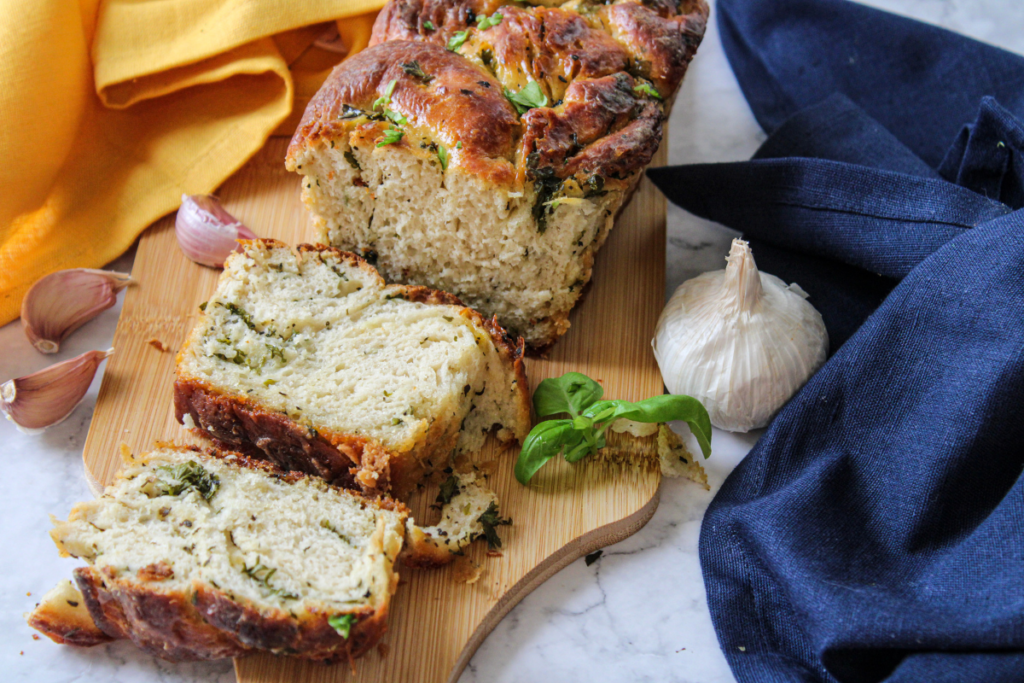 A loaf of savory herb bread on a wooden cutting board with a few slices cut. Fresh basil and garlic bulbs are nearby. A yellow cloth and a blue cloth are placed in the background.