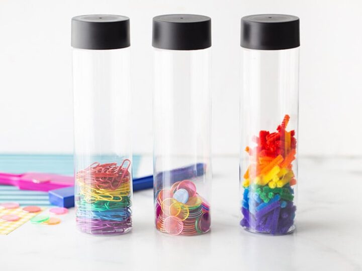Three clear cylindrical containers with black lids hold different colorful office supplies: paperclips, push pins, and binder clips. The background includes colorful stickers and scissors.