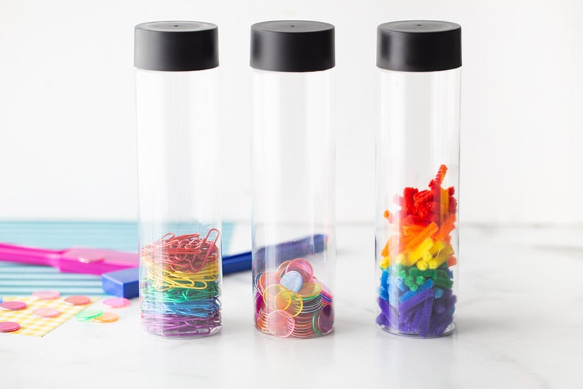 Three clear cylindrical containers with black lids hold different colorful office supplies: paperclips, push pins, and binder clips. The background includes colorful stickers and scissors.