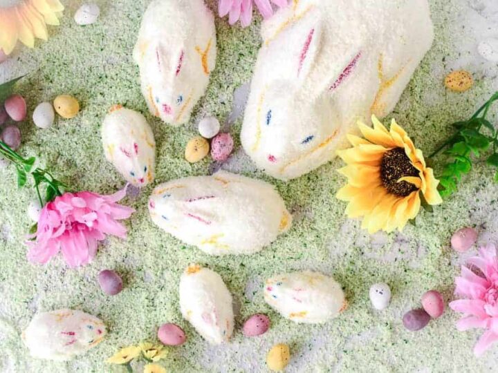 Coconut-covered bunny cakes of various sizes surrounded by colorful candies, artificial flowers, and green-colored shredded coconut, resembling grass. The arrangement is on a white surface, creating a festive spring or Easter theme.