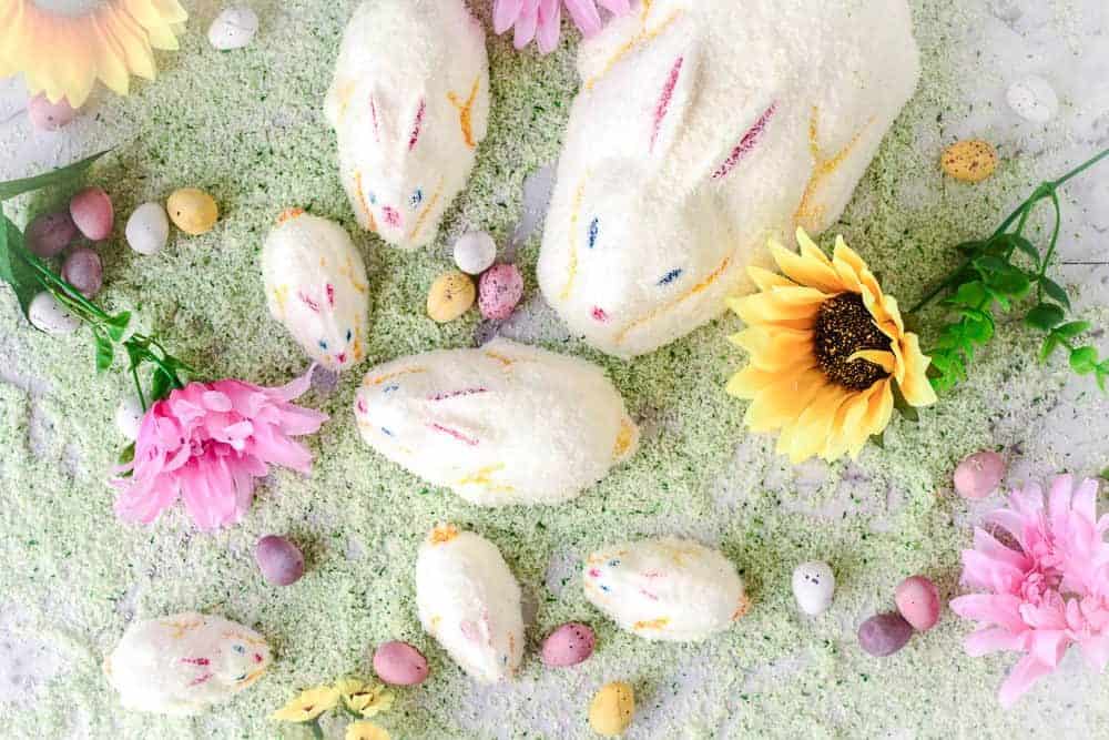 Coconut-covered bunny cakes of various sizes surrounded by colorful candies, artificial flowers, and green-colored shredded coconut, resembling grass. The arrangement is on a white surface, creating a festive spring or Easter theme.