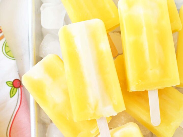 Yellow popsicles with white swirls are arranged on a bed of ice cubes in a metal tray. A colorful, patterned cloth is partially visible on the side.