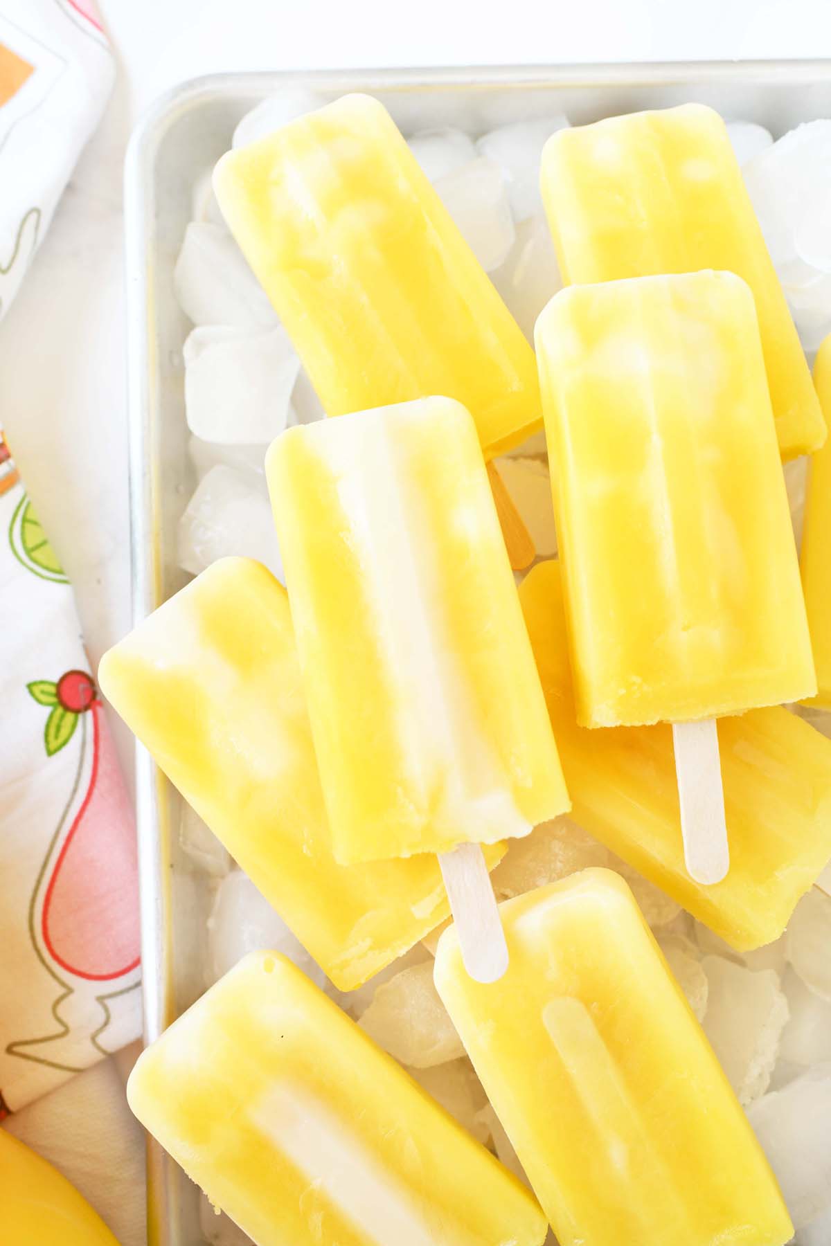 Yellow popsicles with white swirls are arranged on a bed of ice cubes in a metal tray. A colorful, patterned cloth is partially visible on the side.