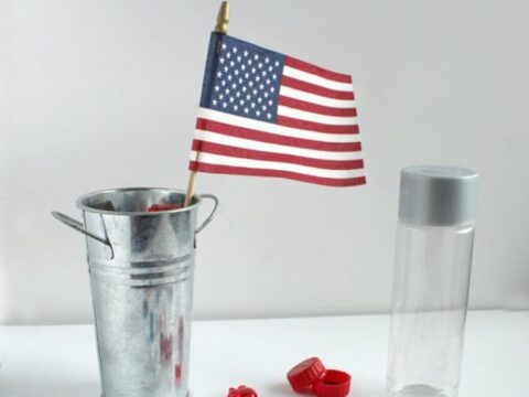 A tin pail holds a small American flag. Surrounding it are a clear plastic bottle, decorative items like letters spelling USA, beads, and miniature red, white, and blue objects, suggesting a patriotic sensory bottle project.