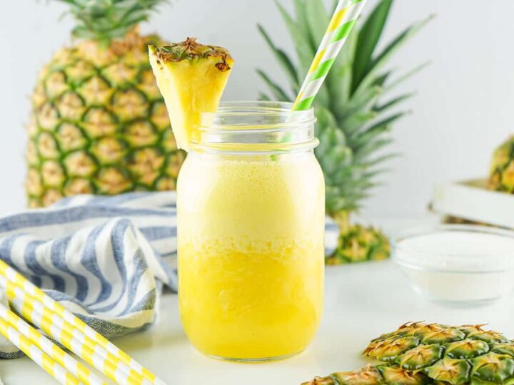 A refreshing pineapple smoothie in a mason jar, garnished with a pineapple wedge and a yellow-and-white striped straw. Whole pineapples and a blue-striped cloth are in the background, with extra straws on the side.