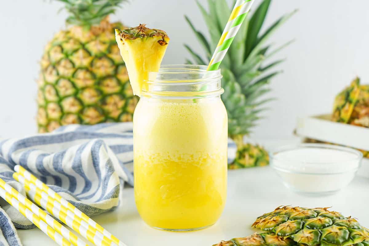 A refreshing pineapple smoothie in a mason jar, garnished with a pineapple wedge and a yellow-and-white striped straw. Whole pineapples and a blue-striped cloth are in the background, with extra straws on the side.