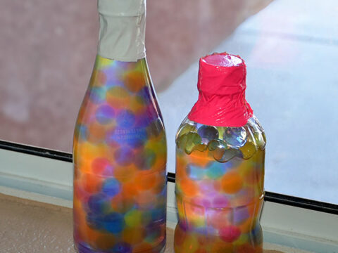 Two clear plastic bottles filled with colorful, water-absorbent beads stand side by side on a windowsill. The taller bottle has a white cap, while the shorter one has a red cap. The sunlight highlights the vibrant colors of the beads.