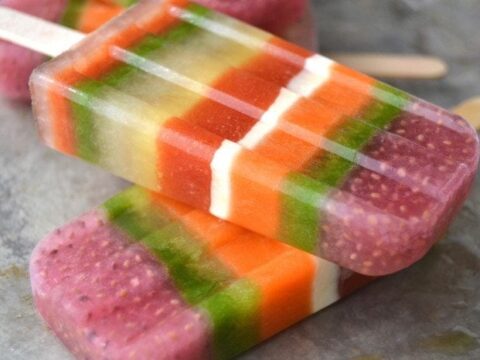 Two colorful layered popsicles with stripes of pink, purple, green, orange, white, and red resting on a gray surface. The vibrant colors hint at various fruity flavors, creating a refreshing summer treat.